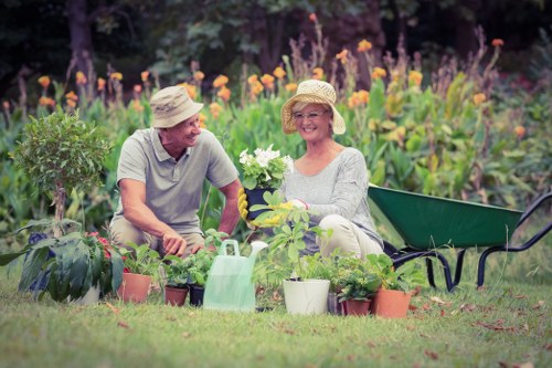 The image shows a mature man and woman sitting together in a well-maintained front garden with lush green grass and vibrant flower beds. They are wearing wide-brimmed straw hats and casual gardening clothes, appearing to enjoy a gardening activity. The woman is holding a white flowering plant, while the man is smiling and engaging with her. Surrounding them are several potted plants, a green watering can, and a black garden bench positioned on the right side of the scene. The background features multicoloured flowers and purple-flowered plants, indicating a blooming garden area, with natural lighting suggesting a clear, sunny day. The scene reflects a tidy outdoor space suitable for gardening and landscaping services, featuring a grassy lawn, flower borders, and garden accessories, typical of a residential environment in Yiewsley, West London.
