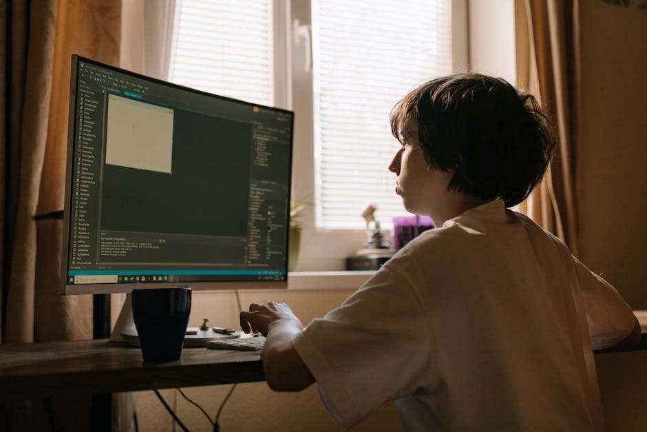 A person with short, dark hair and wearing a white shirt, seated at a wooden desk in a well-lit room, working on a computer with a large monitor displaying an editing or coding interface. The backgrou